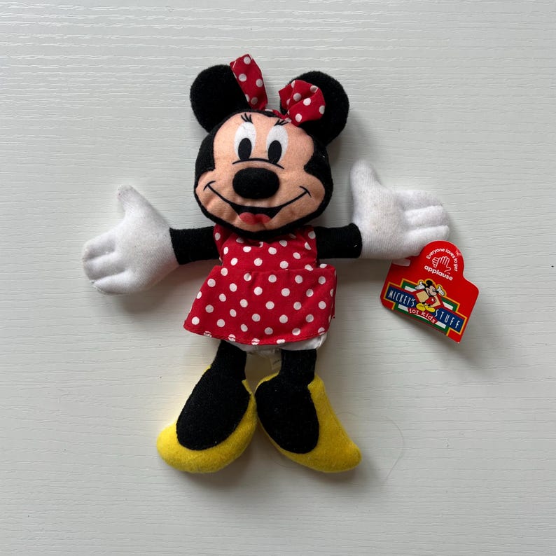 May include: A Minnie Mouse soft toy wearing a red dress with white polka dots and a matching bow. The doll has black ears, arms, and legs, with white gloves and yellow shoes. A red tag with the text "Mickey's Stuff for Kids" is attached.