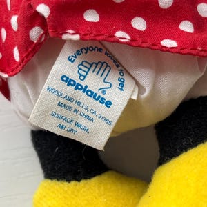 May include: A close-up of a tag from an Applause plush toy. The tag is white with blue text and a logo of two hands. The tag reads "Everyone loves to get applause" and "Made in China". The toy has a red and white polka dot pattern.