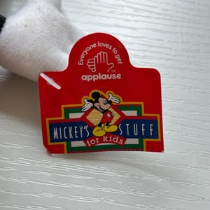 May include: A red tag with the text "Everyone loves to get applause" and "Mickey's Stuff for kids". The tag features a cartoon image of Mickey Mouse. The tag is attached to a white plush toy.