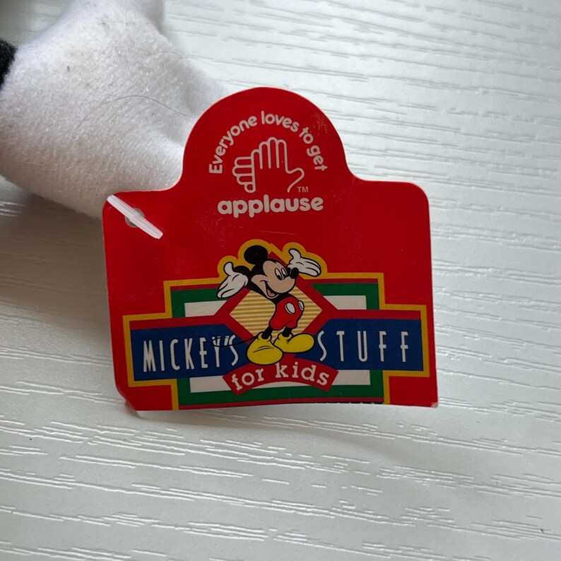 May include: A red tag with the text "Everyone loves to get applause" and "Mickey's Stuff for kids". The tag features a cartoon image of Mickey Mouse. The tag is attached to a white plush toy.