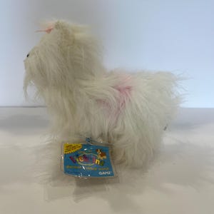 Puede incluir: Un juguete de peluche de perro blanco con pelaje largo y desgreñado y una pequeña mancha rosa en la espalda. Un pequeño lazo rosa está sujeto a la parte superior de la cabeza. Una etiqueta rectangular azul está unida al frente con el texto Webkinz y Ganz.