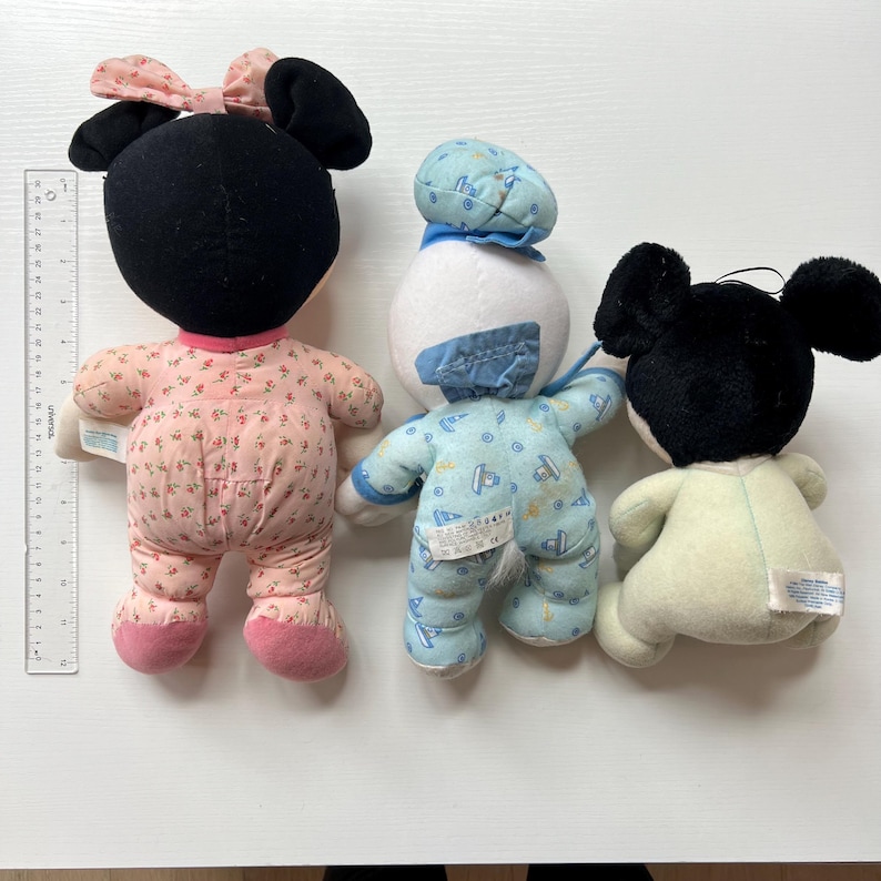 May include: Three vintage plush Disney character dolls. Minnie Mouse in a pink floral print outfit with a bow, Donald Duck in a blue outfit, and Mickey Mouse in a cream-colored outfit. The dolls are soft and appear to be well-loved.