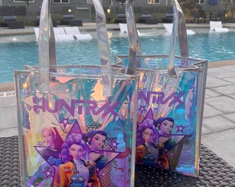 May include: Two clear tote bags with iridescent accents and silver handles. Each bag features a colorful graphic of stylized female figures and the text "HUNTRX" in a bold font. The bags are sitting on a dark surface near a pool.