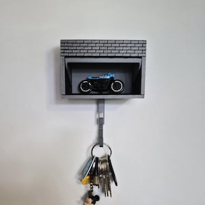 Puede incluir: Un llavero de pared gris con forma de garaje, con una pequeña motocicleta azul en su interior. La parte superior tiene una textura similar a ladrillos. Un juego de llaves cuelga debajo, con un llavero de Mickey Mouse.