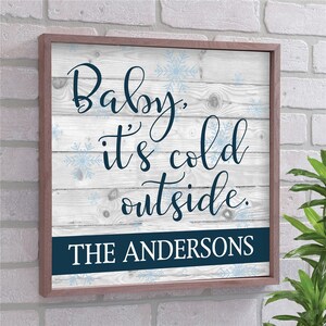 May include: Square wooden framed sign with the text "Baby, it's cold outside." in navy blue script. The background is white with a wood grain pattern and light blue snowflake accents. The bottom has a navy blue banner with the text "THE ANDERSONS."