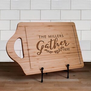 May include: A brown wooden cutting board with a handle on the side. The board is engraved with the text "The Millers Gather Here".