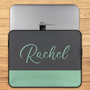 May include: A laptop sleeve with the name "Rachel" in a cursive font, in a light teal color, against a dark gray background. The bottom portion of the sleeve is light teal. The sleeve is on a wooden surface.