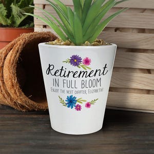 May include: White ceramic plant pot with the text "Retirement IN FULL BLOOM" and floral decorations. The pot contains a green succulent plant. The text "ENJOY THE NEXT CHAPTER, ELIZABETH!" is also included.
