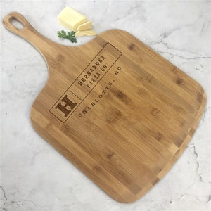 May include: A wooden pizza peel with a handle. The peel is engraved with the text "Hernandez Pizza Co. Charlotte, NC" and the letter "H".