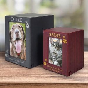 May include: Two wooden pet urns with photo frames. The larger urn is black with the name "DUKE" and the dates "2011-2024" engraved on it. The smaller urn is burgundy with the name "SADIE" and the dates "2009-2023" engraved on it. Both urns have paw prints engraved on them.