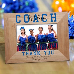 May include: A wooden picture frame with a blue and white inscription that reads "Coach Melissa" and "Thank You" with the text "You were the best coach ever! Cheer Squad 2024". The frame contains a photo of four cheerleaders holding blue pom-poms.