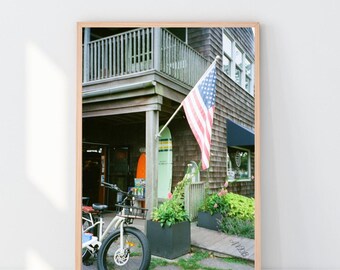 May include: A framed photograph of a building with an American flag waving in the wind. A white bicycle with a black tire is parked in front of the building. Two surfboards are leaning against the building. Green plants are in black planters.