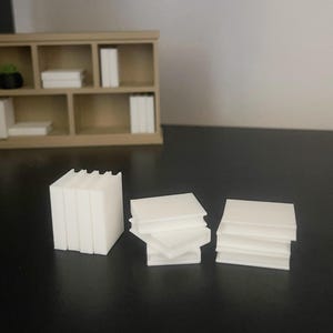 May include: A miniature beige bookshelf with several white books and a small green plant. In the foreground, there are stacks of white square blocks, resembling miniature books. The items are arranged on a dark surface against a neutral wall.