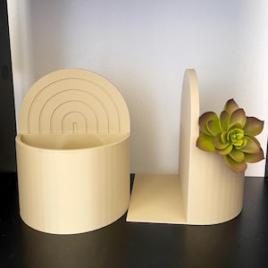 May include: Two beige decorative planters. One is a half-circle with a rainbow design, and the other is a half-circle with a succulent plant. Both planters are on a black surface.