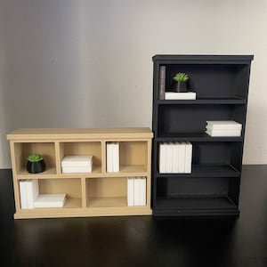 Puede incluir: Se muestran dos estanter&iacute;as en miniatura, una beige y otra negra. La estanter&iacute;a beige tiene m&uacute;ltiples compartimentos con pilas de libros blancos y una peque&ntilde;a planta en maceta. La estanter&iacute;a negra tiene tres estantes con pilas de libros blancos y una peque&ntilde;a planta en maceta.