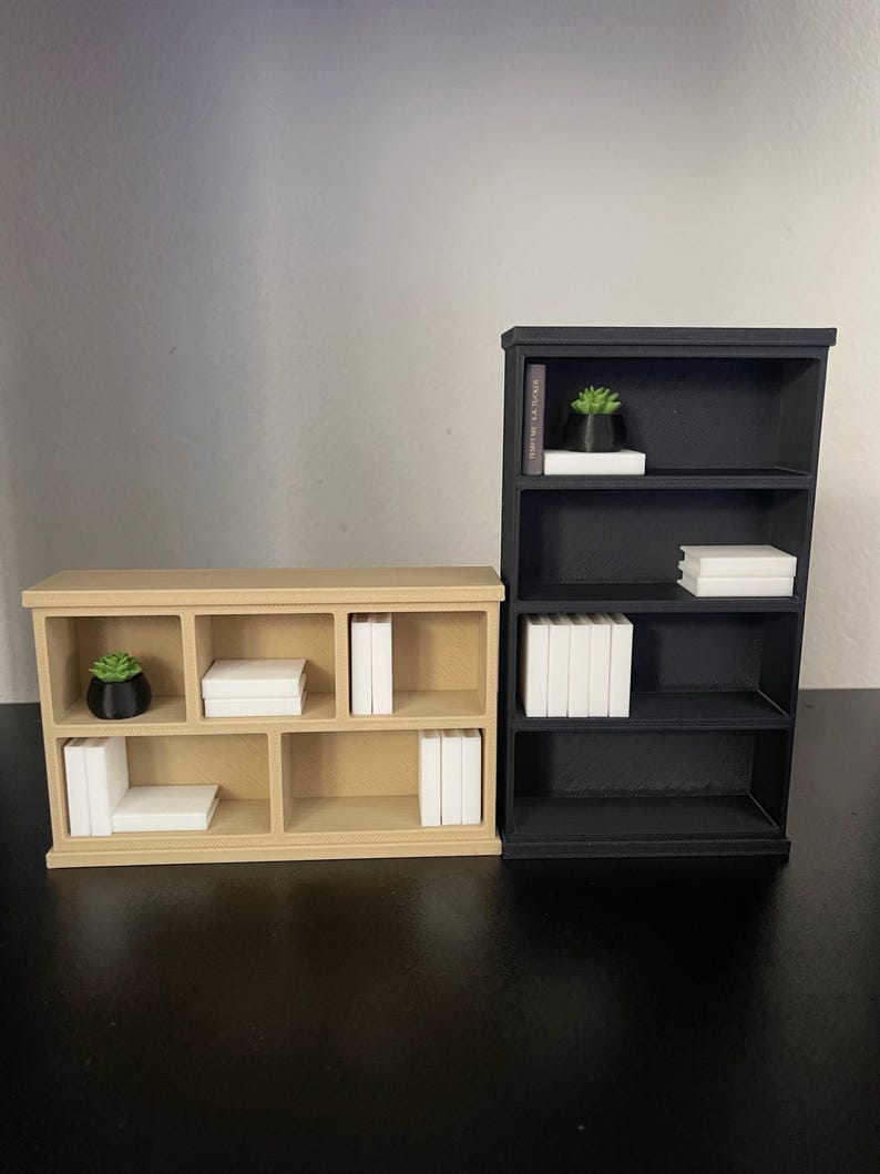 Puede incluir: Se muestran dos estanter&iacute;as en miniatura, una beige y otra negra. La estanter&iacute;a beige tiene m&uacute;ltiples compartimentos con pilas de libros blancos y una peque&ntilde;a planta en maceta. La estanter&iacute;a negra tiene tres estantes con pilas de libros blancos y una peque&ntilde;a planta en maceta.