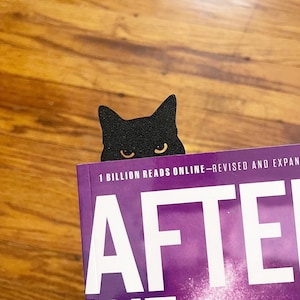 May include: A black cat-shaped bookmark peeks over the top of a book. The book has a purple cover with the title "AFTER WE" in large white letters. The text "1 BILLION READS ONLINE" is also visible.