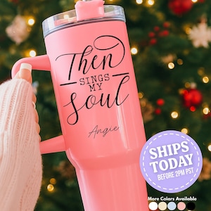May include: A pink insulated tumbler with a handle, featuring the text "Then Sings My Soul" and the name "Angie" in black script. The tumbler has a clear lid and a straw. Additional colors are available.