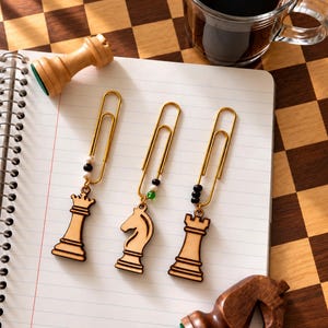 May include: Three gold-toned metal paperclips, each featuring a wooden chess piece charm. The charms represent a queen, a knight, and a rook. Each paperclip is accented with small, colourful beads in black, white, and green, attached to the top.