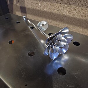 May include: A handcrafted metal flower sculpture. The flower petals are made from spoons, with a threaded rod stem and a washer detail. The sculpture is silver and sits on a metal surface.