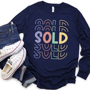 May include: Navy blue long-sleeve shirt with a colorful "SOLD" graphic. The shirt is paired with blue jeans and white sneakers.