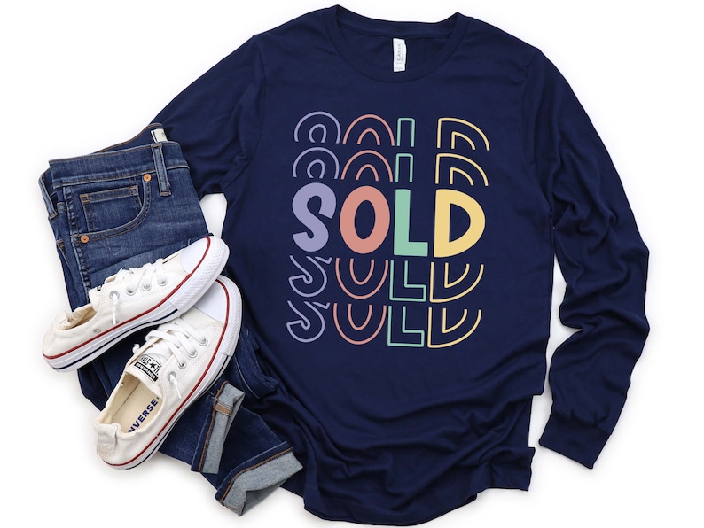 May include: Navy blue long-sleeve shirt with a colorful "SOLD" graphic. The shirt is paired with blue jeans and white sneakers.