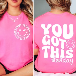 Pu&ograve; includere: T-shirt rosa acceso con testo bianco e design a faccina sorridente. Davanti: "Don't Stress, Do Your Best, Rock the Test." Dietro: "You Got This" con l'hashtag #testday.