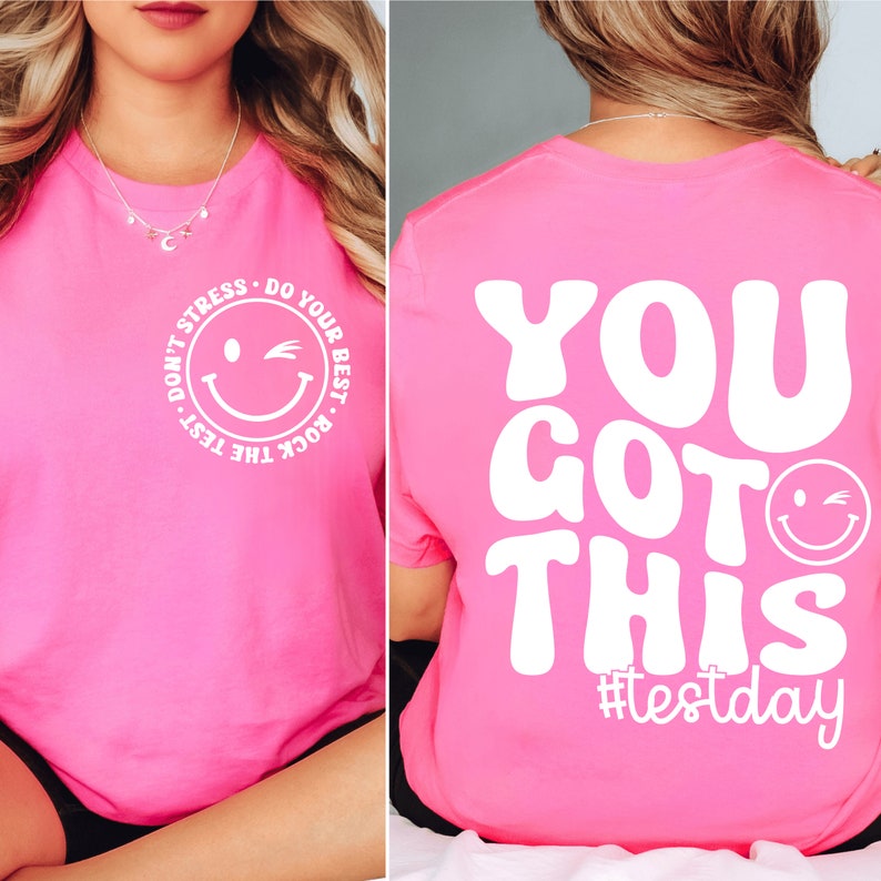 Pu&ograve; includere: T-shirt rosa acceso con testo bianco e design a faccina sorridente. Davanti: "Don't Stress, Do Your Best, Rock the Test." Dietro: "You Got This" con l'hashtag #testday.
