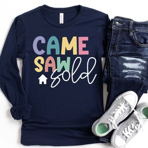 May include: Navy blue long-sleeve shirt with the text "Came Saw Sold" in colorful letters and a small house icon. The shirt is paired with blue jeans and gray sneakers.
