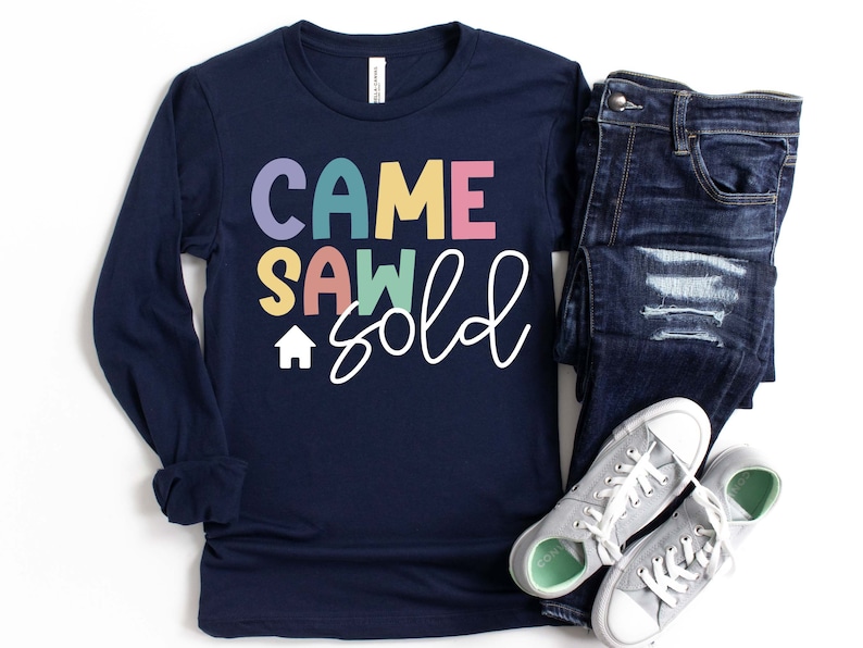 May include: Navy blue long-sleeve shirt with the text "Came Saw Sold" in colorful letters and a small house icon. The shirt is paired with blue jeans and gray sneakers.