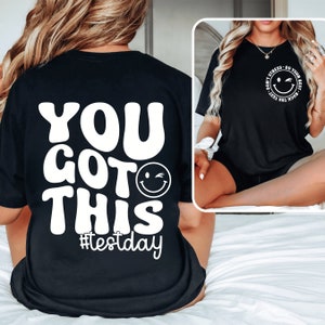 Pu&ograve; includere: T-shirt nero con testo bianco e una faccina sorridente che fa l'occhiolino. La parte posteriore della maglietta dice "YOU GOT THIS #testday" in un font retr&ograve;. La parte anteriore della maglietta ha un piccolo disegno circolare con una faccina sorridente e le parole "Don't Stress - Do Your Best - Rock The Test".
