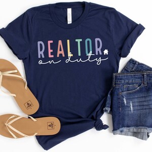 May include: A navy blue t-shirt with the text "REALTOR on duty" in rainbow colors. The shirt is paired with denim shorts and gold sandals.