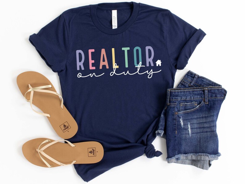 May include: A navy blue t-shirt with the text "REALTOR on duty" in rainbow colors. The shirt is paired with denim shorts and gold sandals.