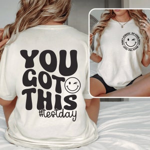 Pu&ograve; includere: T-shirt color crema con la scritta "YOU GOT THIS" in grandi caratteri neri in stile retr&ograve;. Sotto il testo c'&egrave; una faccina sorridente che fa l'occhiolino, con l'hashtag "#testday" sotto. La maglietta presenta anche un disegno pi&ugrave; piccolo con una faccina sorridente che fa l'occhiolino e la scritta "DON'T STRESS - DO YOUR BEST - ROCK THE TEST".