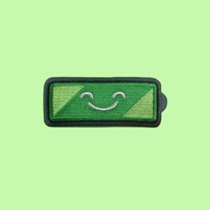 May include: A rectangular embroidered patch featuring a battery icon design. The patch has a black border and a green center with a lighter green diagonal section on the left and right. A white stitched smiling face is centered on the battery.