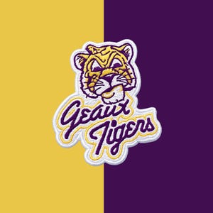 May include: An embroidered patch featuring a stylized tiger head in yellow and purple thread with white outlines. Below the tiger head, the text Geaux Tigers is stitched in a purple cursive font with a yellow outline on a white background.