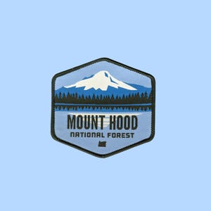 Puede incluir: Un parche bordado hexagonal que muestra un pico de montaña blanco cubierto de nieve sobre un bosque oscuro y un lago azul. El texto MOUNT HOOD NATIONAL FOREST está impreso en letras negras, con un pequeño contorno del estado de Oregon abajo.