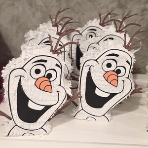 Puede incluir: Se muestran varias pi&ntilde;atas blancas con forma de Olaf, con contornos negros, grandes sonrisas y narices de zanahoria. Las pi&ntilde;atas tienen cuernos marrones en forma de ramita y est&aacute;n cubiertas de flecos blancos. Est&aacute;n dispuestas sobre una superficie blanca.