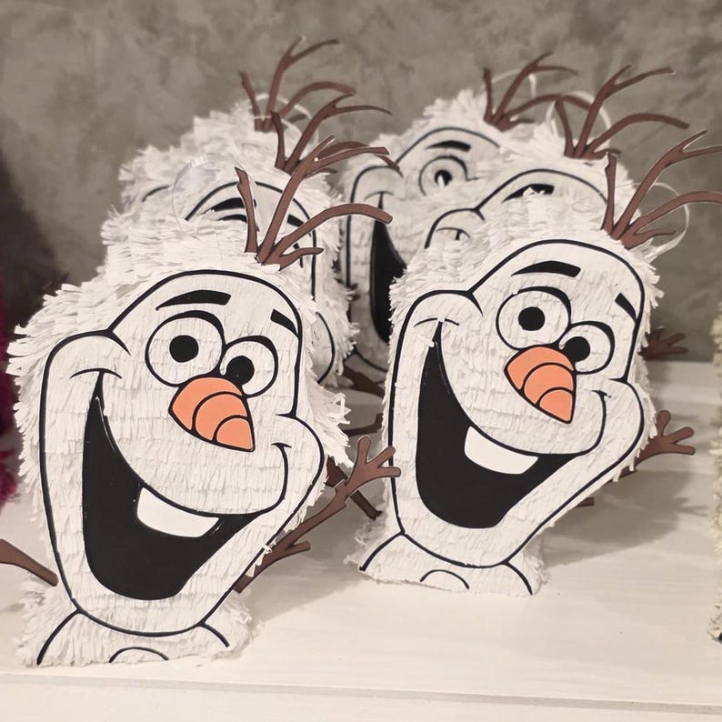 Puede incluir: Se muestran varias pi&ntilde;atas blancas con forma de Olaf, con contornos negros, grandes sonrisas y narices de zanahoria. Las pi&ntilde;atas tienen cuernos marrones en forma de ramita y est&aacute;n cubiertas de flecos blancos. Est&aacute;n dispuestas sobre una superficie blanca.