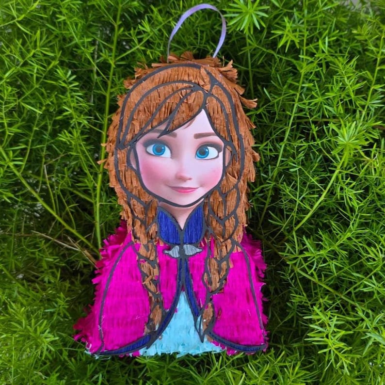 Puede incluir: Una pi&ntilde;ata hecha a mano con la forma de un personaje de dibujos animados, con una cara sonriente, ojos azules, cabello casta&ntilde;o trenzado y un vestido rosa y azul. Se adjunta una cinta morada para colgar.