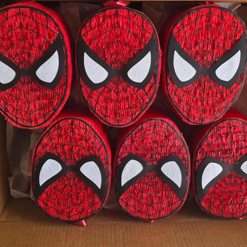 Op de afbeelding: Zes rode Spider-Man gezicht pi&ntilde;ata's met zwarte oogcontouren en witte oogvormen zijn gerangschikt in een kartonnen doos. De pi&ntilde;ata's hebben een textuur met een webachtig patroon.