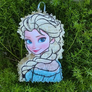 Puede incluir: Una pi&ntilde;ata con forma de personaje de dibujos animados con pelo rubio y un vestido azul. La pi&ntilde;ata tiene una franja blanca alrededor de la cabeza y una cinta azul para colgarla. El personaje tiene ojos azules y una ligera sonrisa.