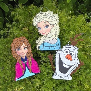 May include: Three character-shaped pinatas featuring Elsa with blonde hair and a blue dress, Anna with braided brown hair and a pink cape, and a white snowman with a carrot nose and stick arms. Each item has a fringed paper texture.