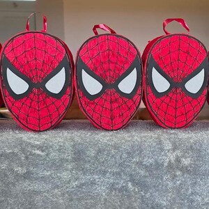 Op de afbeelding: Drie rode Spider-Man-rugzakken met zwarte oogmaskers en witte oogvormen. De rugzakken hebben een webpatroon en rode handvatten. De tassen worden op een grijze ondergrond getoond.