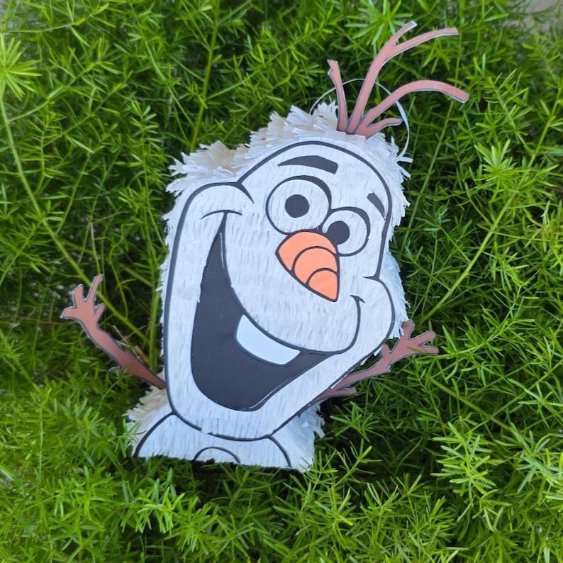 Puede incluir: Una pi&ntilde;ata blanca de Olaf con una gran boca negra, una nariz de zanahoria naranja y ojos negros. La pi&ntilde;ata tiene brazos de palo marrones y una cabeza blanca y esponjosa. El fondo es un lecho de follaje verde.