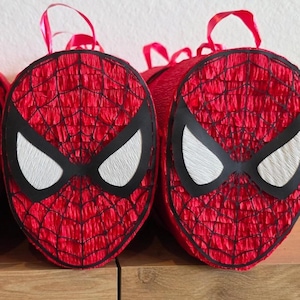 Op de afbeelding: Rode en zwarte Spider-Man gezicht pi&ntilde;atas met witte oogdetails en rode lint handvatten. De pi&ntilde;atas zijn gemaakt van rood cr&ecirc;pepapier en zwarte accenten, perfect voor een superhelden themafeest.