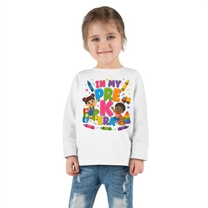 Op de afbeelding: Een wit shirt met lange mouwen en een kleurrijke afbeelding met de tekst IN MY PRE K ERA. Het ontwerp bevat getekende kinderen, alfabetblokken, een speelgoedvrachtwagen, een stapeltoren van ringen en diverse kleurpotloden eromheen.