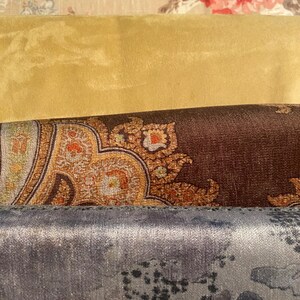 May include: A close-up of several fabric swatches. The top swatch is beige with a floral pattern in red and pink. Below is a solid gold fabric, followed by a brown paisley design. The bottom swatch is a gray-blue with a mottled pattern.