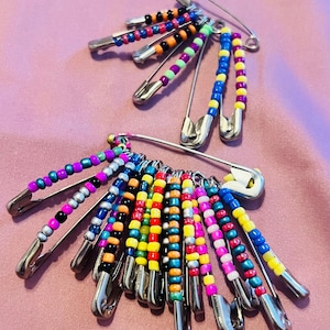 May include: Two sets of beaded safety pins. Each pin is adorned with colorful beads in various hues, including blue, pink, yellow, and green. The pins are arranged on a pink surface, creating a vibrant and eye-catching display.