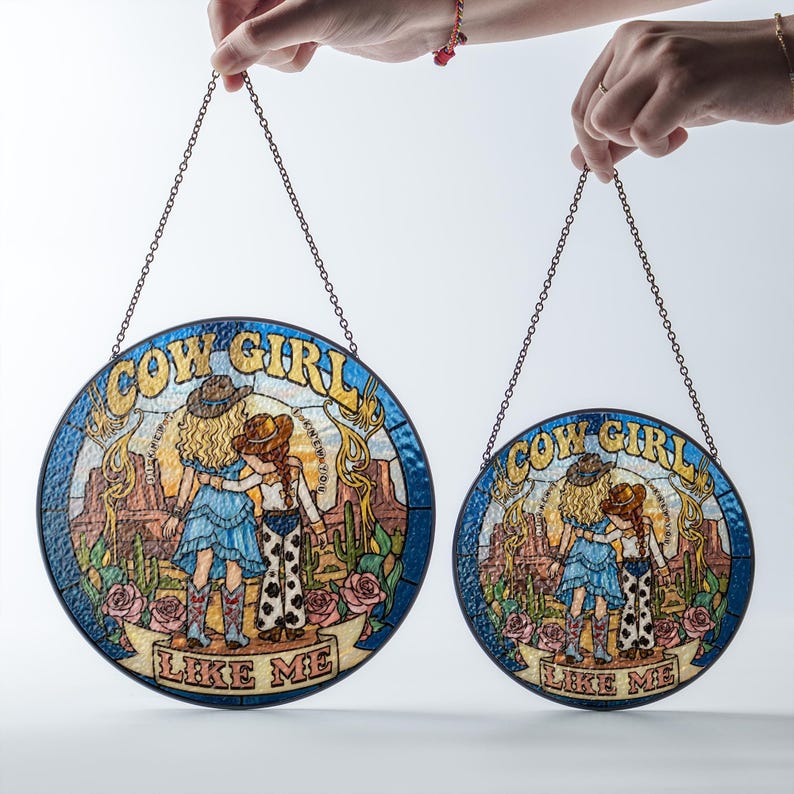 May include: Two circular stained glass suncatchers with metal chains, featuring a desert scene with two figures in cowboy hats, boots, and western attire. The text COW GIRL LIKE ME is printed in blue and red on a banner at the bottom of each piece.